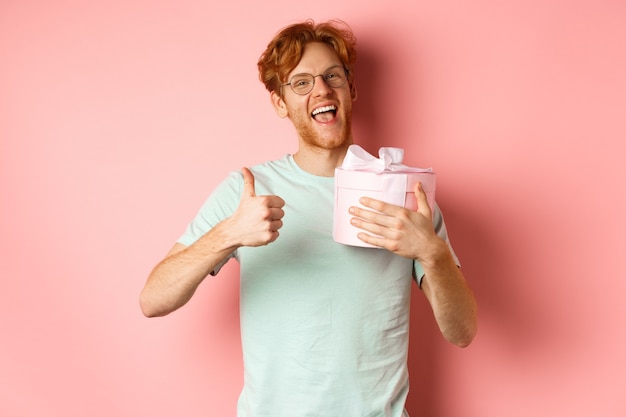 Valentines day and romance concept. cheerful young man holding box with gift and showing thumbs-up, thanking for present, standing over pink background.