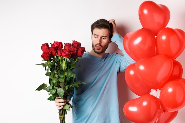 발렌타인 데이 로맨스. 혼란스러운 남자친구는 머리를 긁적이며 데이트를 위해 빨간 장미 꽃다발을 보고 있습니다. 우유부단한 느낌의 꽃과 풍선을 가진 남자, 흰색 배경