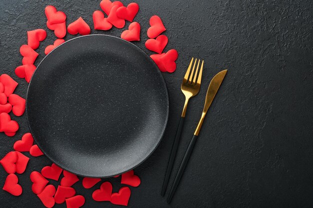 Valentines day romantic table setting Empty or closeup of a dinner black plate knife fork and decorative silk hearts on black background Holiday concept Copy space for inscriptions