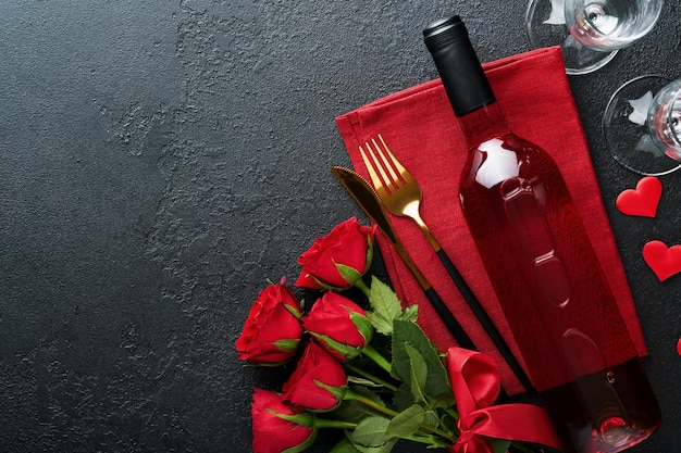 Valentines day romantic table setting Empty closeup of red roses wine candles dinner black plate knife fork and decorative silk hearts on black background Holiday concept Copy space Top view