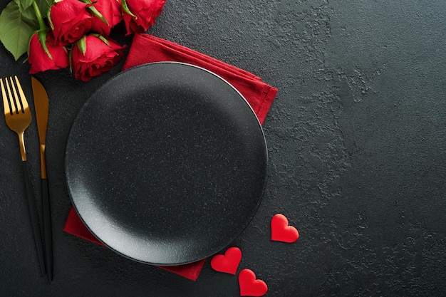 Valentines day romantic table setting Empty closeup of red roses wine candles dinner black plate knife fork and decorative silk hearts on black background Holiday concept Copy space Top view