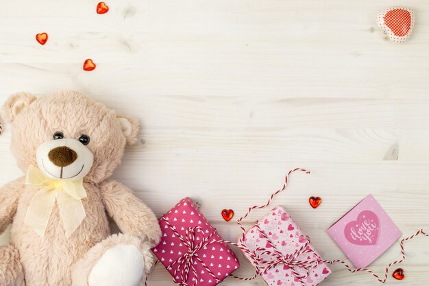 Valentines Day scene with teddy bear, gift boxes, greeting card and hearts on a light wooden background.