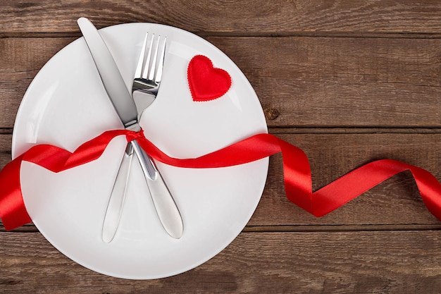 Сервировка стола на день святого валентина с тарелкой, вилкой, ножом, красным сердцем и ленточным фоном