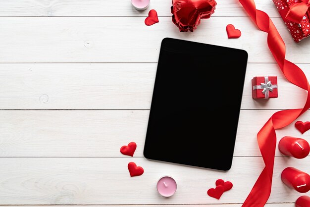 Valentines Day. Tablet with black screen with valentine decorations candles, balloons and confetti top view on white wooden background