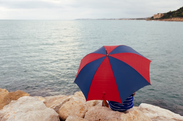 Van achter anonieme man zittend op de rotsen met een paraplu aan zee terwijl het regent