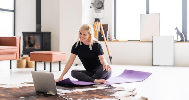 Van achteren gezien fitte sportvrouw in fitnesskleding in de moderne woonkamer met behulp van online fitnesstrainingsprogramma in laptop terwijl ze op fitnessmat ligt.