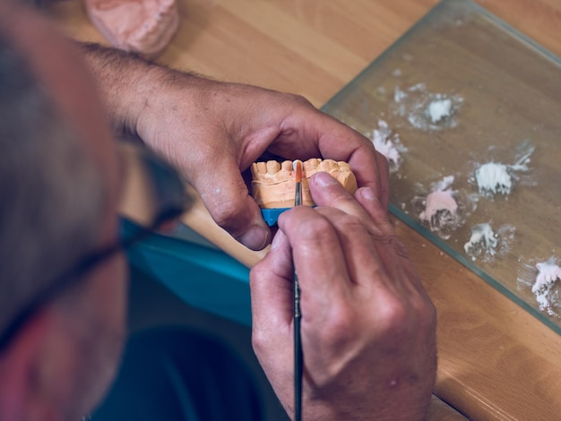 Foto van bovenaf een onherkenbare tandtechnicus die een borstel gebruikt voor het bedekken van een kunstgebit met keramiek in de werkplaats