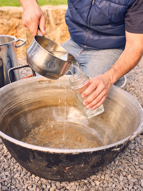 Van bovenaf van het gewas, onherkenbare man die water uit de kookpot in een glazen pot giet over een metalen bassin dat op steenachtige grond is geplaatst