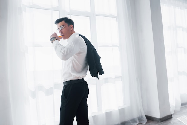 Van de achterkant. Luxe uitziende man in klassieke kleding staat in de kamer en houdt de bovenkant van het pak en glas vast met alcohol.