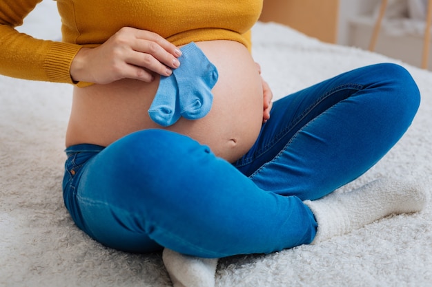 Van mij met liefde. ontspannen vrouwelijke persoon die haar buik aanraakt en kleine sokken vasthoudt terwijl ze op het bed zit