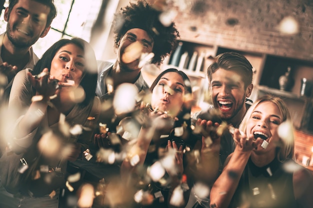 Foto vang het plezier! vrolijke jonge mensen die confetti blazen en glimlachen terwijl ze samen genieten van een feestje