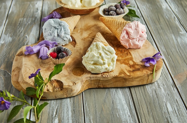 Vanilla, blueberry and raspberry ice cream in waffle cones on wooden board with berries and flowers.