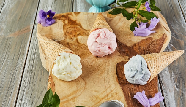 Vanilla, blueberry and raspberry ice cream in waffle cones on wooden board with berries and flowers.