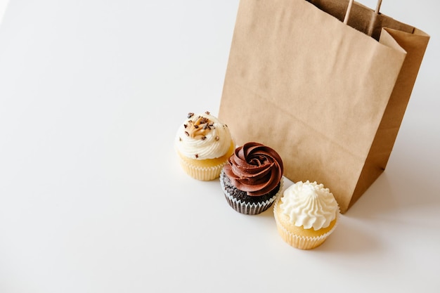 Photo vanilla chocolate and caramel cupcakes and brown paper bag on white table background