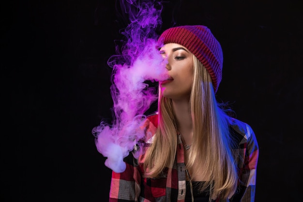 Photo vaping girl. young hipster woman vape e-cig on studio on black background. hip-hop style. close up.