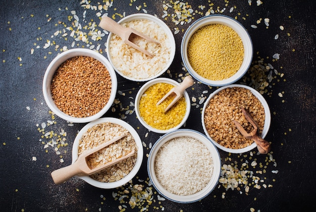 Variety of cereal flakes rice, millet, buckwheat, oatmeal. Superfood in white ceramic bowls on dark old concrete