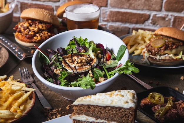 Varietà di piatti, insalata con formaggio di capra hamburger fatti in casa e patatine fritte, ali di pollo con jalapeños drink e torta sul tavolo di legno. immagine isolata.