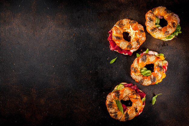 Variety of homemade bagels sandwiches with sesame and poppy seeds, cream cheese,  ham, radish, arugula, cherry tomatoes, cucumbers, dark concrete background copy space above