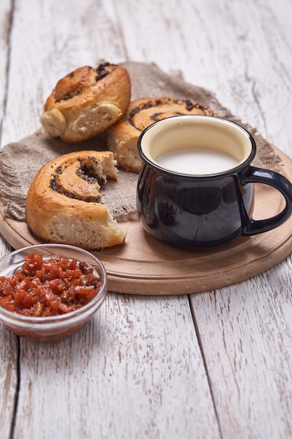 Variety of homemade puff pastry buns cinnamon served with milk cup, jam, butter as breakfast over white plank wooden table