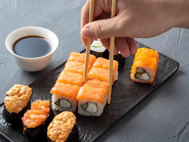 A variety of Japanese rolls on a black plate with soy and a hand picking one up with chopsticks