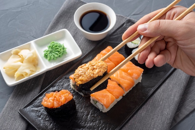 Разнообразные японские роллы и суши на текстурированной черной тарелке. Вид сбоку. Бамбуковые палочки удерживают один гункан. Закрыть вверх