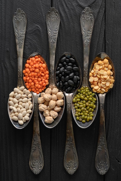 Variety of legumes in old silver spoons on a black wooden\
background