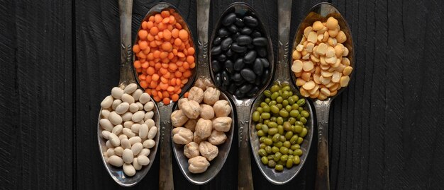 Variety of legumes in old silver spoons on a black wooden background