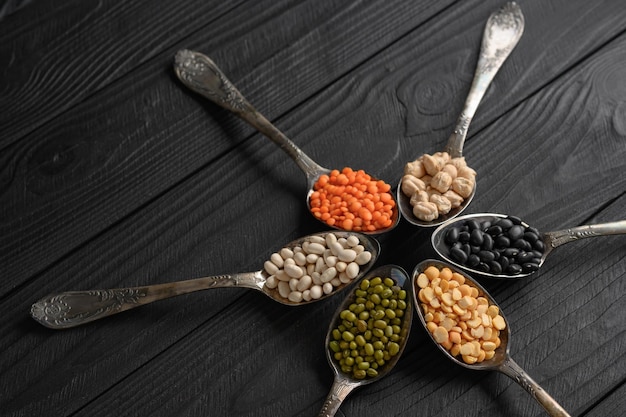 Variety of legumes in old silver spoons on a black wooden
background