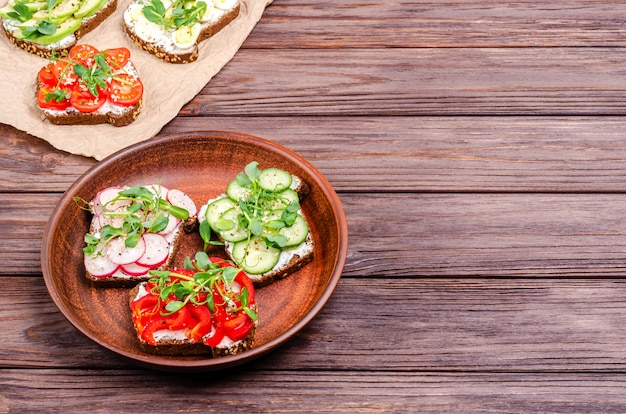 A variety of mini sandwiches with cream cheese and vegetables on a clay grater and paper on a wooden surface