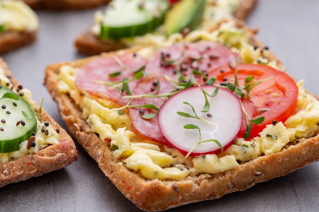 Variety of mini sandwiches with cream cheese, vegetables and salami. Sandwiches with cucumber, radish, tomatoes, salami on a gray background, top view. Flat lay.