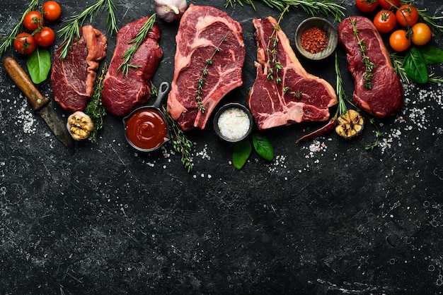 Variety of raw aged beef steaks tbone tomahawk striploin tenderloin new york steak for grilling with spices on stone background On a black stone background