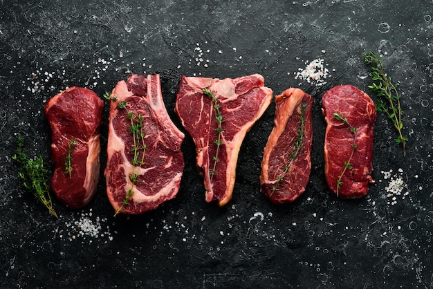 A variety of raw beef steaks for grilling with seasonings Tbone striploin steak ribeye Top view On a black stone background