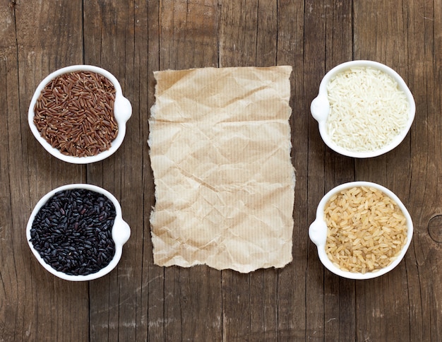 Variety of rice in bowls around old paper on wooden table top view