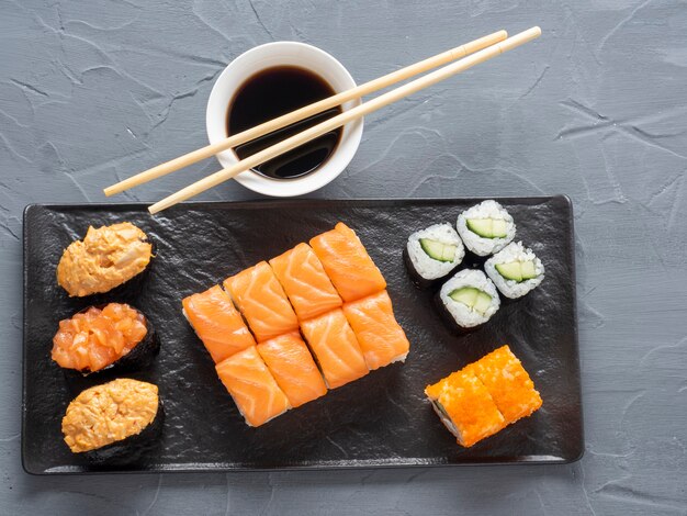 A variety of rolls and sushi gunkan nested on a black plate. next to it are bamboo wasabi sticks and sauce