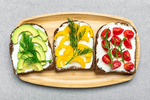 Variety of sandwiches for breakfast - slice of whole grain dark\
bread, yellow bell pepper, cream cheese, avocado, cherry tomatoes,\
garnished with dill, green arugula on table top view flat lay