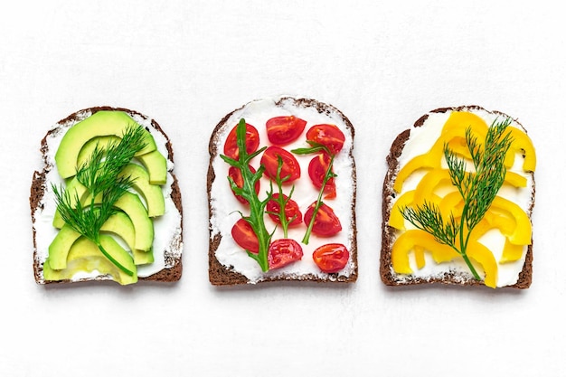 Photo variety of sandwiches for breakfast - slice of whole grain dark bread, yellow bell pepper, cream cheese, avocado, cherry tomatoes, garnished with dill, green arugula on white table top view flat lay