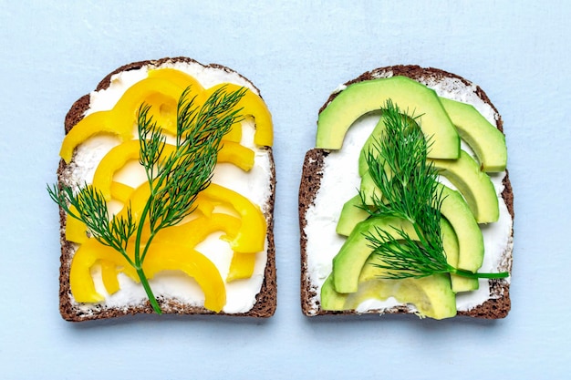 Varietà di panini per colazione - fetta di pane scuro integrale, peperone giallo, crema di formaggio, avocado guarnito con aneto verde su tavolo blu vista dall'alto piatto concetto di cibo vegano