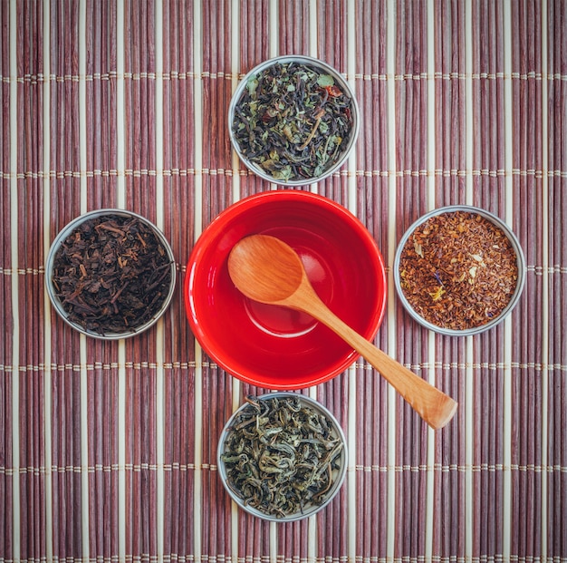 Photo a variety of teas and a wooden spoon in an empty red cup on a bamboo mat