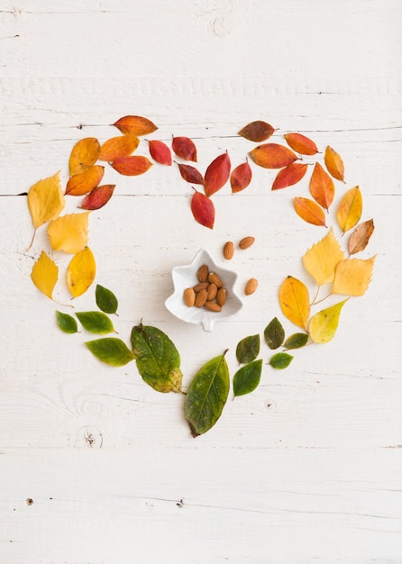 ハート型のさまざまな秋の色とりどりの葉と白い木製の背景にアーモンドナッツとボウル