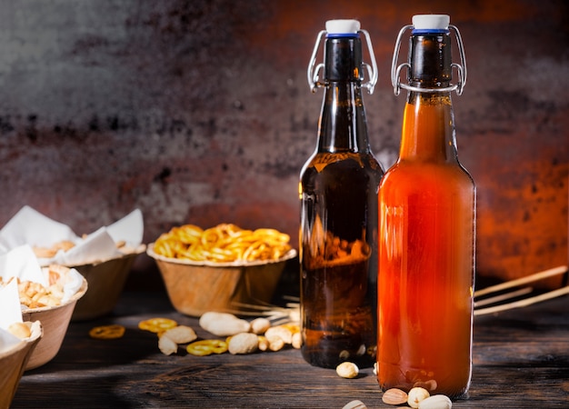 Various beer snacks in plates like pistachios, small pretzels and peanuts near two bottles of filtered and unfiltered beer on dark wooden desk. Food and beverages concept