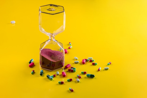 Various capsules and tablets laid out on a yellow  next to which are an hourglass with red grit.Health .