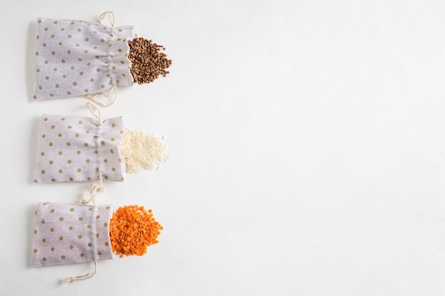 Various cereals buckwheat, rice and lentils in canvas bags scattered on the white concrete background. Flat lay with place for text.
