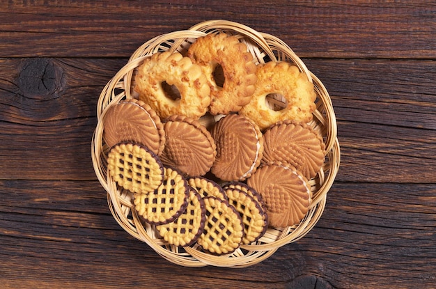 Various cookies in wicker plate on dark wooden table, top view