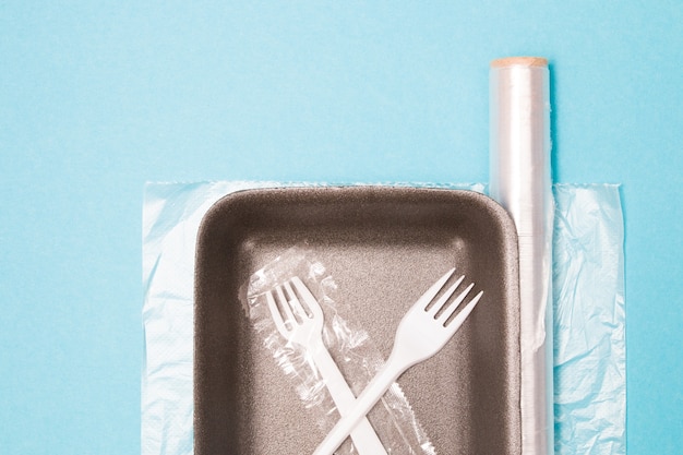 Various disposable plastic packaging on a blue background, a food tray, a roll of plastic tape and a bag for freezing food, a disposable fork and a drinking tube, environmental pollution concept