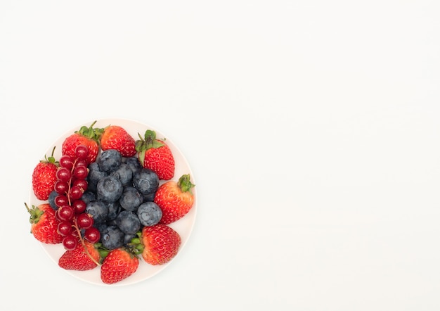 Various fresh summer berries strawberries, currant, blueberries on white plate on white background.