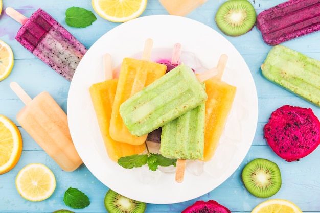 Various fruit popsicles are placed on the blue wooden board background, kiwi popsicles, orange popsicles, dragon fruit popsicles, cantaloupe popsicle