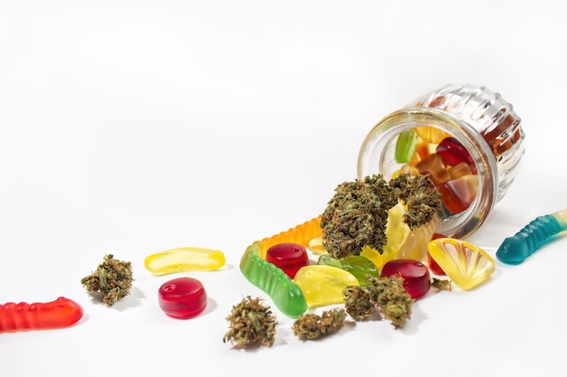 Various gummies and dried medical marijuana buds fell out of the embossed glass jar On a white background Lots of empty space