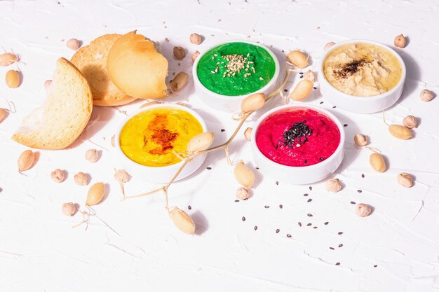 Various hummus dips, the flat lay of hummus in different colors with spinach, beetroot, turmeric and vegetables, vegan snack. White putty background, copy space
