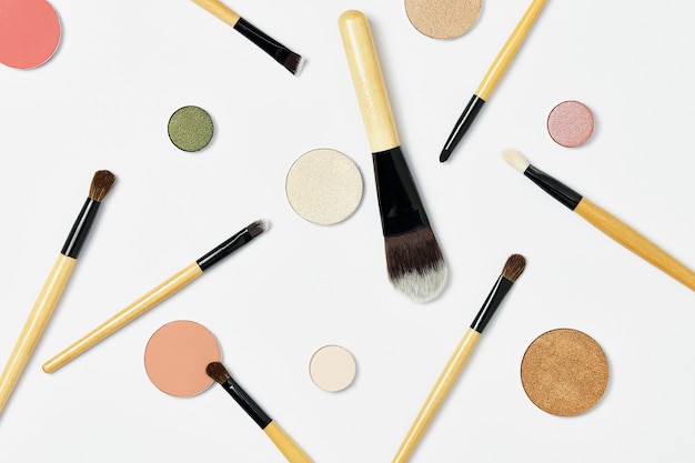 Various professional makeup brushes with a wooden brown handle lie randomly on a white background, around a lot of different multi-colored round eye shadow palettes for makeup. Top view, flatlay.