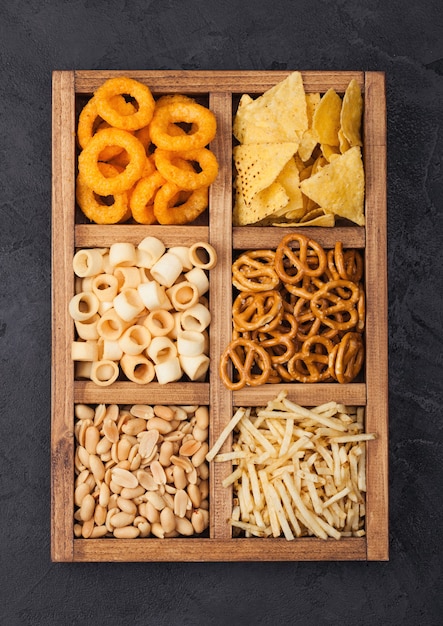 Various snacks in vintage wooden box on black kitchen background. Onion rings,nachos, salty peanuts with potato sticks and pretzels. Suitable for beer and fizzy drinks.